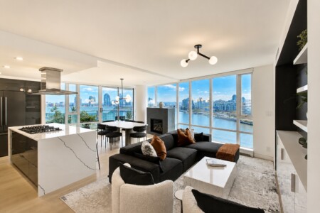Open-plan living area with marble kitchen island, dining table, and a black sectional sofa, overlooking a river and city skyline through large windows.