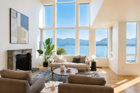 Bright living room with a large window wall showing a lake and distant mountains, a beige sofa and armchairs, and a stone fireplace.