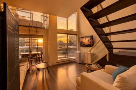 Loft living room at sunset with large windows, wooden staircase, chalkboard wall, and beige sofa facing a TV.