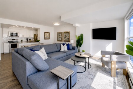 Open-concept living room with a blue sectional sofa, round coffee tables, and a wall-mounted TV in view of a bright kitchen area.