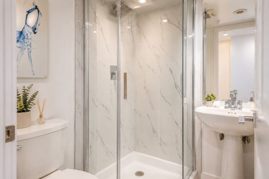 Modern bathroom with a glass-enclosed shower, marble walls, a white toilet, and a pedestal sink with a small plant on the tank area.