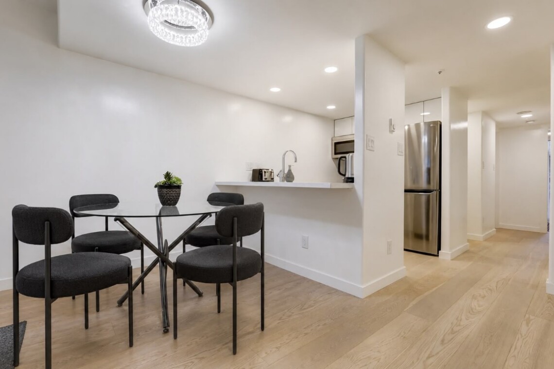 Open-concept kitchen and dining area with glass round table, four dark chairs, white walls, and hardwood floors; stainless fridge and small appliances visible.