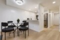 Open-concept kitchen and dining area with glass round table, four dark chairs, white walls, and hardwood floors; stainless fridge and small appliances visible.