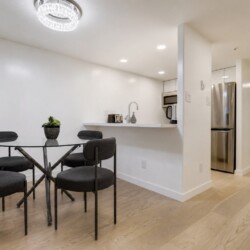 Open-concept kitchen and dining area with glass round table, four dark chairs, white walls, and hardwood floors; stainless fridge and small appliances visible.