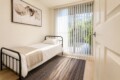 Small bedroom with a single metal framed bed, white linens, and a beige throw blanket by a large glass door with vertical blinds