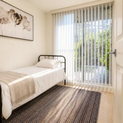 Small bedroom with a single metal framed bed, white linens, and a beige throw blanket by a large glass door with vertical blinds