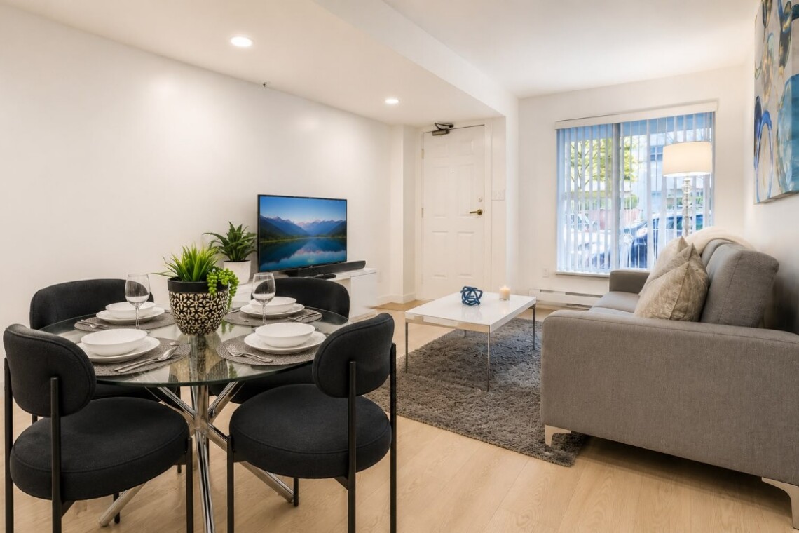 Living room with gray sofa, coffee table, and rug facing a TV; dining area with round glass table and black chairs nearby.