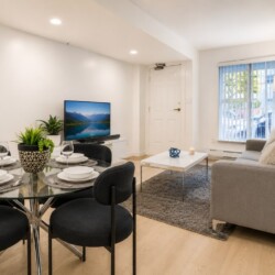 Living room with gray sofa, coffee table, and rug facing a TV; dining area with round glass table and black chairs nearby.