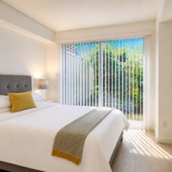 Bright modern bedroom with a white-velvet bed, gray tufted headboard, yellow throw pillow, and striped gray throw; large glass doors with vertical blinds open to a garden.