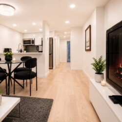 Open-concept living space with a lit fireplace on the right, glass dining table, black chairs, and a modern kitchenette in the background.