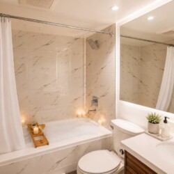 Modern bathroom with marble-tiled bath area, white shower curtain, and wood bath tray holding candles and small plant.