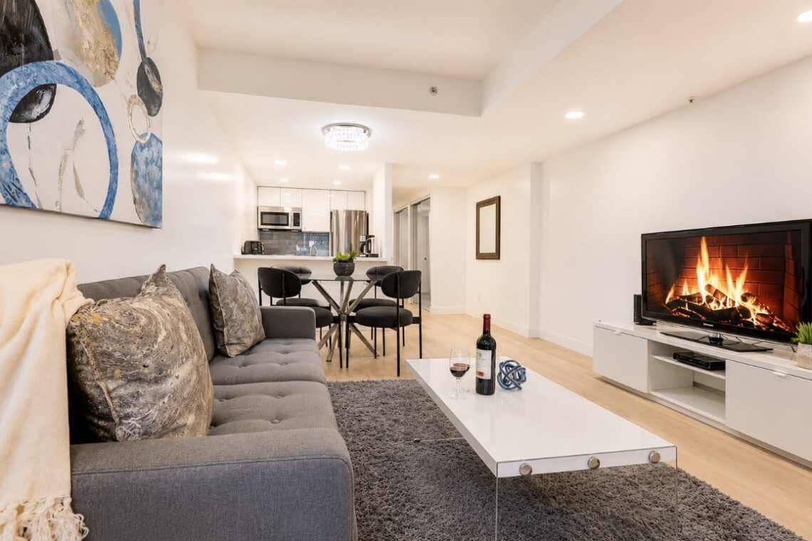 Open-plan living room with a gray sectional sofa, white coffee table, shag rug, and a TV stand with a lit fireplace in the background and dining area beyond.