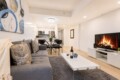 Open-plan living room with a gray sectional sofa, white coffee table, shag rug, and a TV stand with a lit fireplace in the background and dining area beyond.