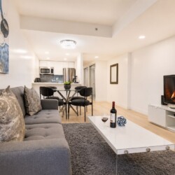 Open-plan living room with a gray sectional sofa, white coffee table, shag rug, and a TV stand with a lit fireplace in the background and dining area beyond.