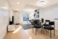 Bright open-plan living and dining area: gray sofa, glass dining table with four black chairs, abstract wall art, and a large window bringing in light.