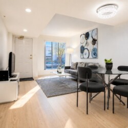 Bright open-plan living and dining area: gray sofa, glass dining table with four black chairs, abstract wall art, and a large window bringing in light.