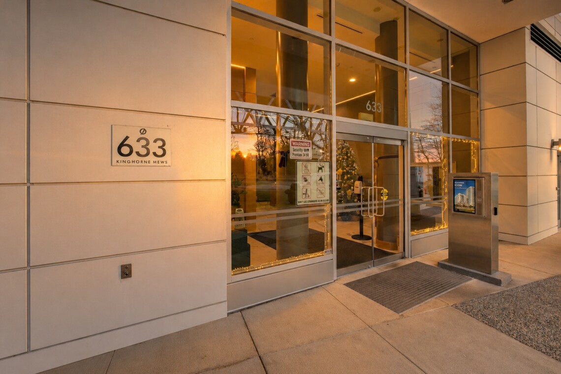 Exterior view of a modern building entrance with glass doors and a 633 Kinghorne Mews sign on beige walls, holiday lights around the doorway, and a metal kiosk nearby.