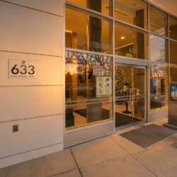 Exterior view of a modern building entrance with glass doors and a 633 Kinghorne Mews sign on beige walls, holiday lights around the doorway, and a metal kiosk nearby.
