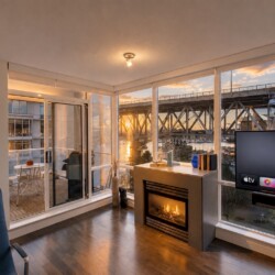 Modern living room with a lit fireplace, flat-screen TV, and floor-to-ceiling windows showing a sunset over a bridge and marina.