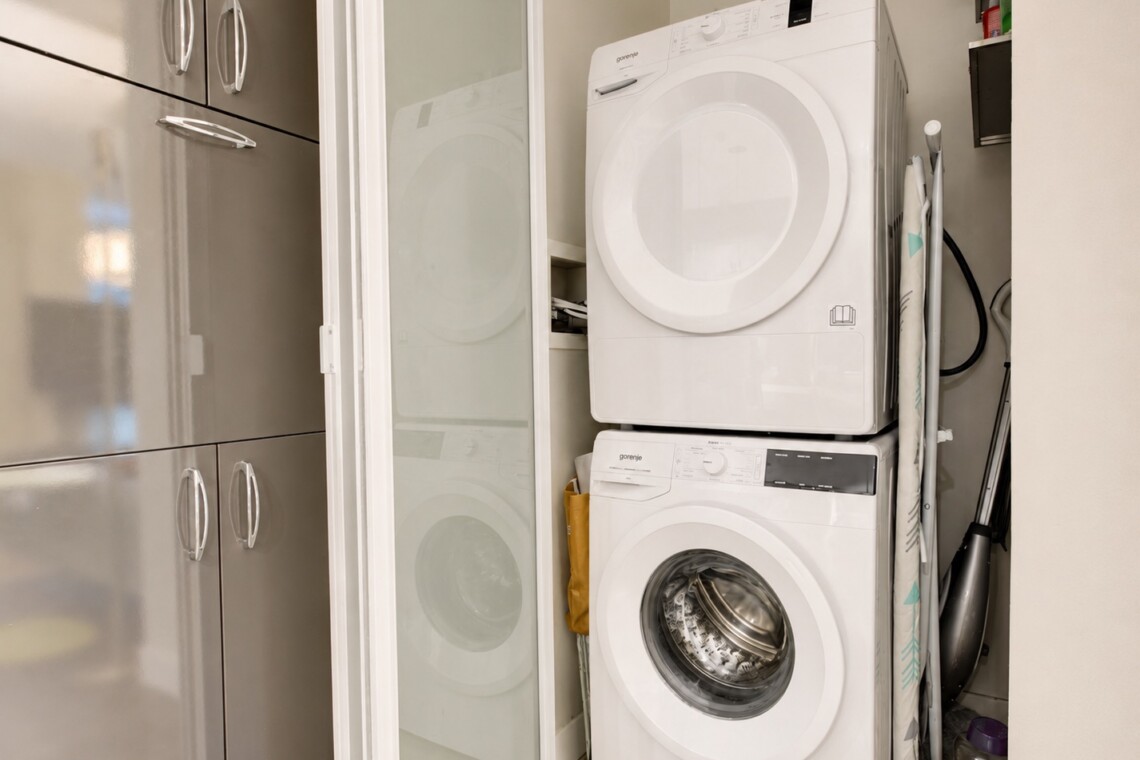 Stacked white front-loading washer and dryer in a narrow laundry closet beside frosted glass doors and tall cabinets with silver handles, plus ironing board nearby.