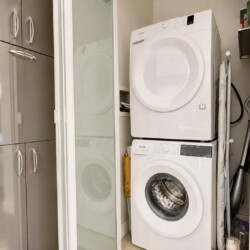 Stacked white front-loading washer and dryer in a narrow laundry closet beside frosted glass doors and tall cabinets with silver handles, plus ironing board nearby.