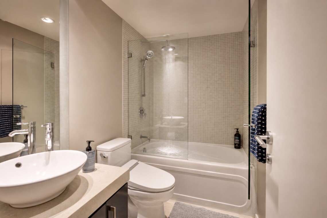 Contemporary bathroom with a glass shower enclosure, white bathtub, and a rounded white basin atop a beige countertop.