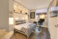 Modern studio bedroom with white built-in shelving, a bed with gray striped bedding, and a glass desk by a large window.