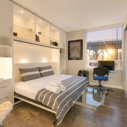 Modern studio bedroom with white built-in shelving, a bed with gray striped bedding, and a glass desk by a large window.