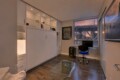 Modern home office with white built-in cabinets, glass coffee table, and a glass desk near a window showing a sunset cityscape and bridge.