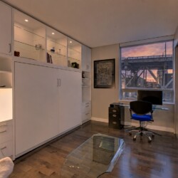 Modern home office with white built-in cabinets, glass coffee table, and a glass desk near a window showing a sunset cityscape and bridge.