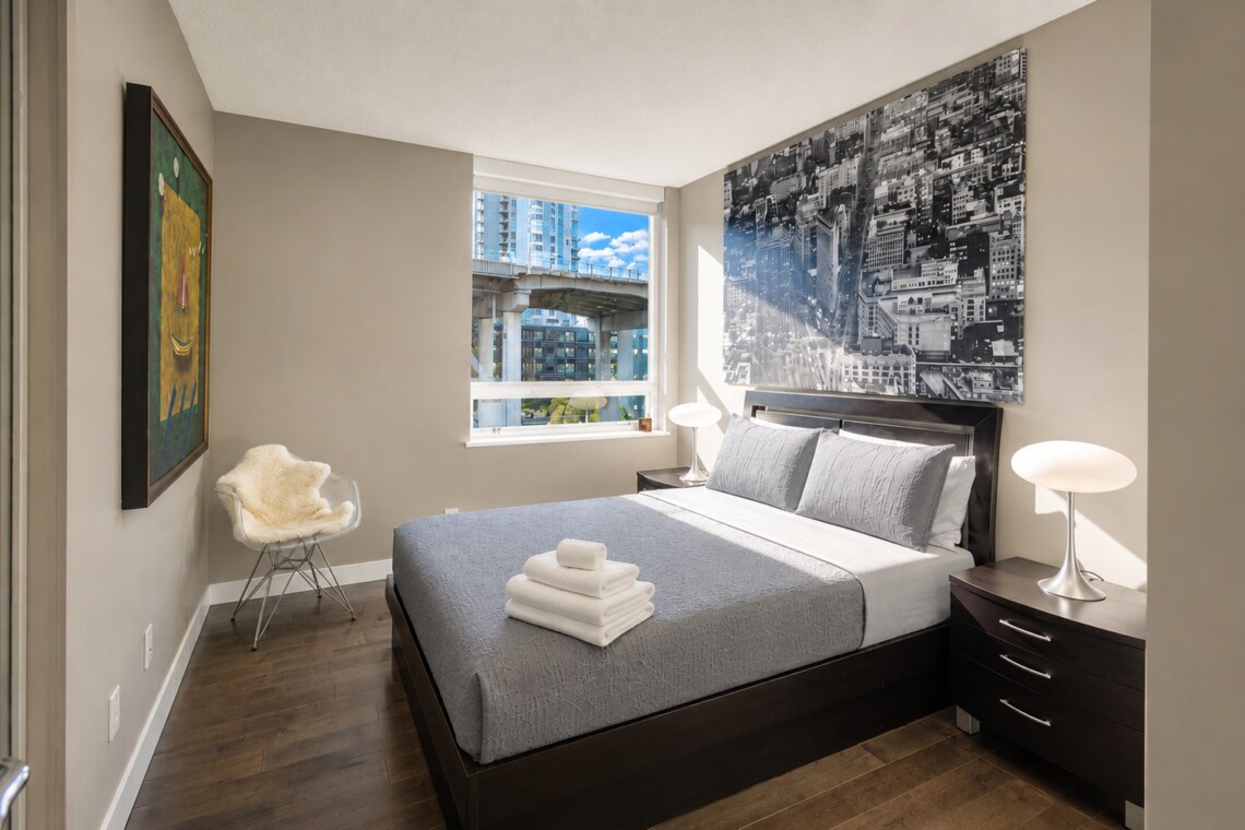 Modern hotel bedroom with a dark wooden bed, gray quilt, and matching pillows; nightstands with lamps, large cityscape artwork above the headboard, and a window view of buildings outside.