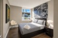 Modern hotel bedroom with a dark wooden bed, gray quilt, and matching pillows; nightstands with lamps, large cityscape artwork above the headboard, and a window view of buildings outside.