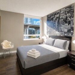 Modern hotel bedroom with a dark wooden bed, gray quilt, and matching pillows; nightstands with lamps, large cityscape artwork above the headboard, and a window view of buildings outside.