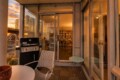 Sunset-lit balcony with a Weber grill, metal chair, and glass doors opening to a book-filled living room.