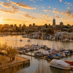 Sunset over a marina with boats and a city skyline, warm orange glow reflecting on the water.