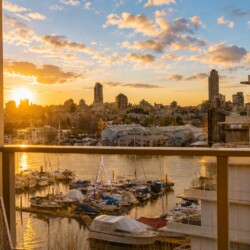 Sunset view over a marina with moored boats and a city skyline, seen from a balcony railing.