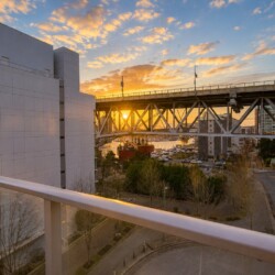 Sunset over a marina and city viewed from a balcony, with a steel bridge, boats, and modern buildings nearby.