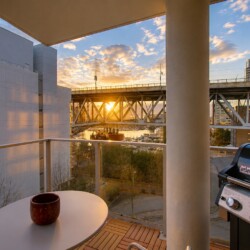 Balcony scene with a round table and red mug, a Weber grill on the right, and a sunset over a bridge and marina in the distance.