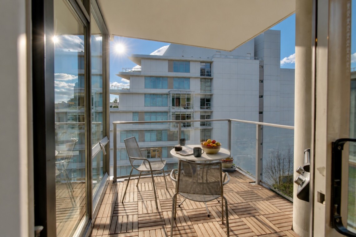 Sunlit balcony with a round table and metal chairs, bowl of fruit and mugs, overlooking modern white apartment buildings.
