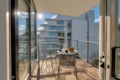 Sunlit balcony with a round table and metal chairs, bowl of fruit and mugs, overlooking modern white apartment buildings.