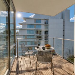 Sunlit balcony with a round table and metal chairs, bowl of fruit and mugs, overlooking modern white apartment buildings.