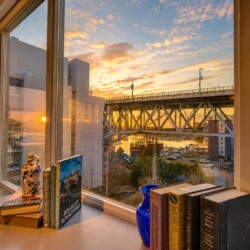 Sunset view through a large window of a bridge over a harbor, with a desk in the foreground holding books and a blue vase.