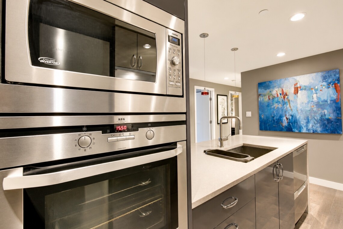 Modern kitchen with stainless steel built-in microwave and double ovens, gray cabinetry, and an island with a sink.