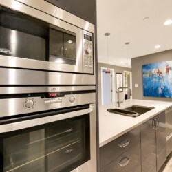 Modern kitchen with stainless steel built-in microwave and double ovens, gray cabinetry, and an island with a sink.