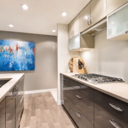 Modern L-shaped kitchen with gray lower cabinets, white countertops, and a gas cooktop under frosted glass cabinets; abstract blue painting on gray wall