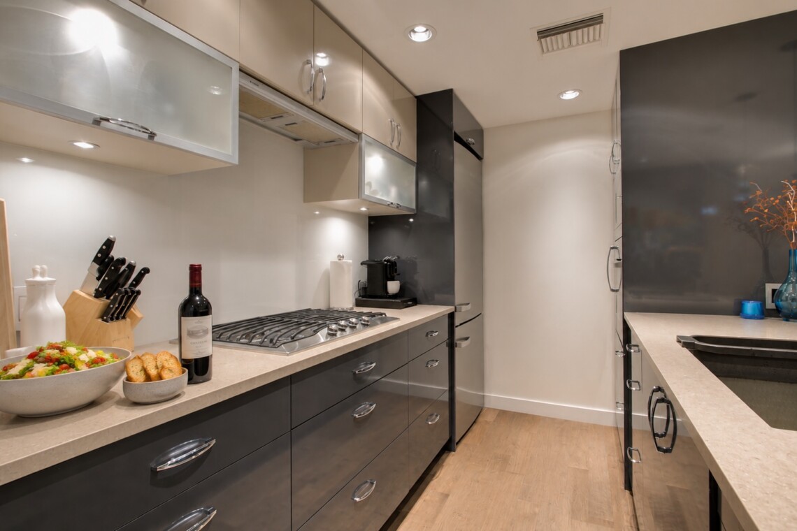 Modern L-shaped kitchen with gray lower cabinets, beige countertops, and a gas range; bowls of salad and bread with a wine bottle on the counter.