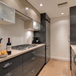 Modern L-shaped kitchen with gray lower cabinets, beige countertops, and a gas range; bowls of salad and bread with a wine bottle on the counter.