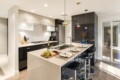 Modern kitchen with a large island, sink, and four black bar stools; fruit board, champagne bottle, glasses on the counter, and pendant lights overhead.