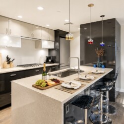 Modern kitchen with a large island, sink, and four black bar stools; fruit board, champagne bottle, glasses on the counter, and pendant lights overhead.