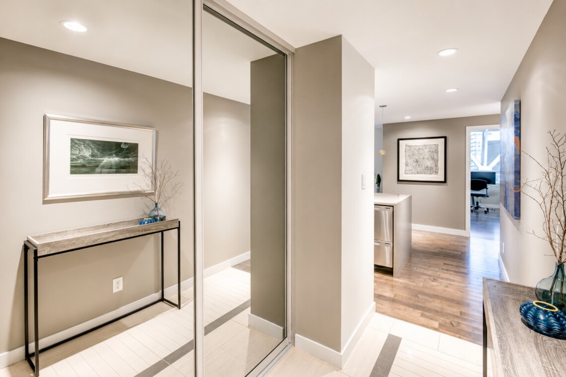Modern hallway with a console table, framed artwork, and a blue vase with bare branches; sliding glass doors reflect the entryway.
